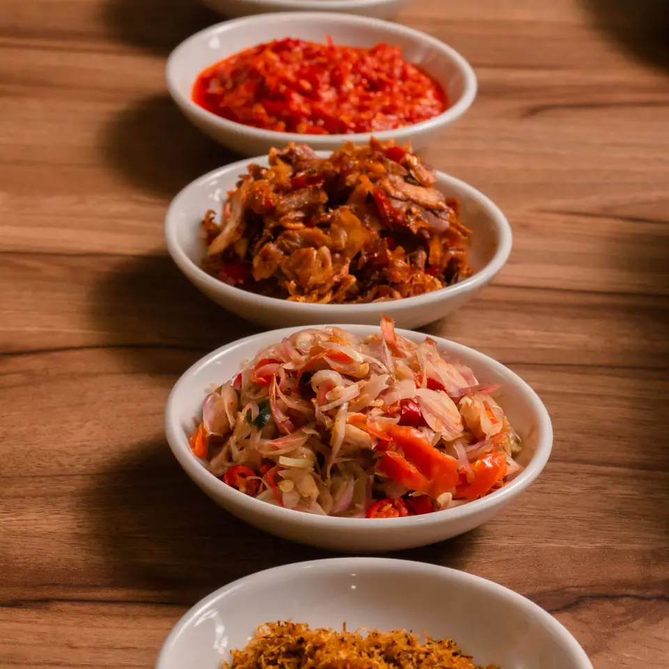 Lima mangkuk sambal berbeda warna berjajar di meja kayu