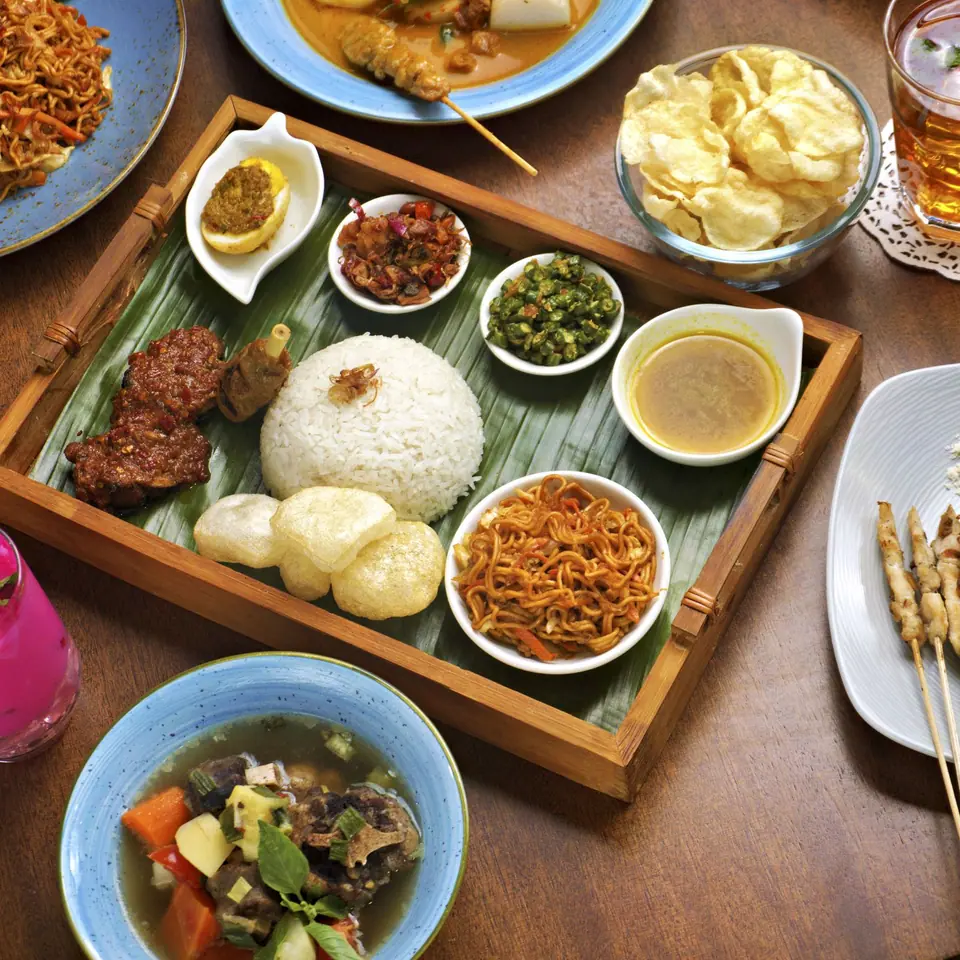 Meja makan dilihat dari atas: nasi di atas daun pisang, sate, sambal, dan es teh